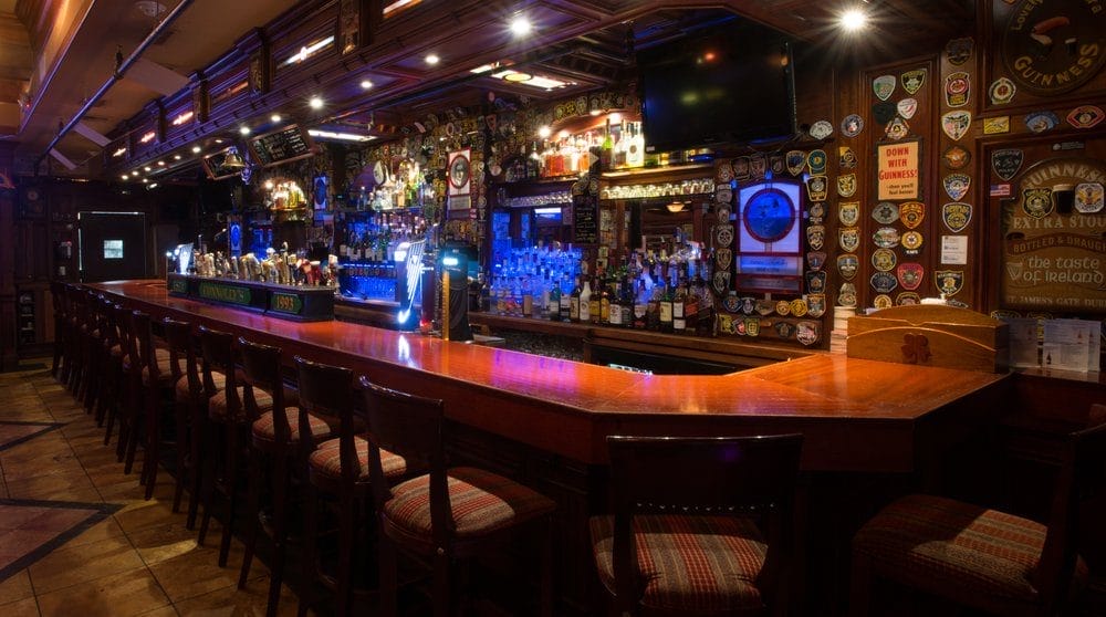 Interior view of an ornate Irish pub featuring a well-lit bar lined with various taps and a wide selection of spirits. The bar is adorned with Irish and Guinness-related memorabilia, including plaques and banners, enhancing the traditional vibe. Stools are arranged neatly, inviting guests to enjoy a drink in this classic setting.