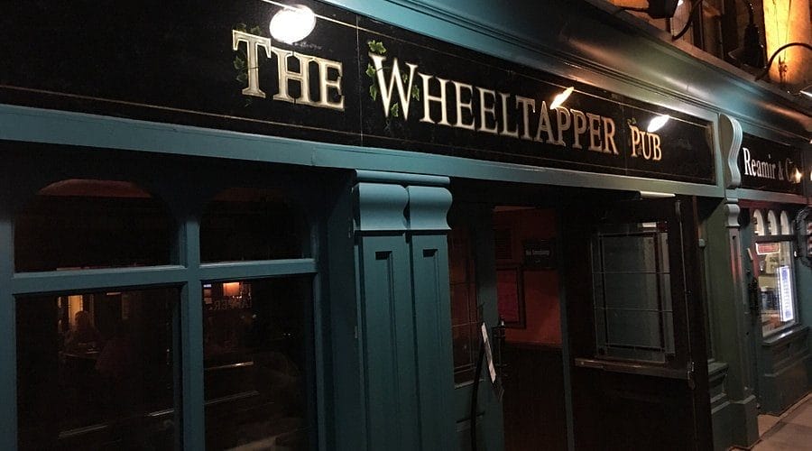 Exterior night view of The Wheeltapper Pub, showcasing its distinctive teal and dark blue façade with the pub's name illuminated in elegant script above the entrance. The welcoming, traditional design hints at a classic Irish pub atmosphere inside.