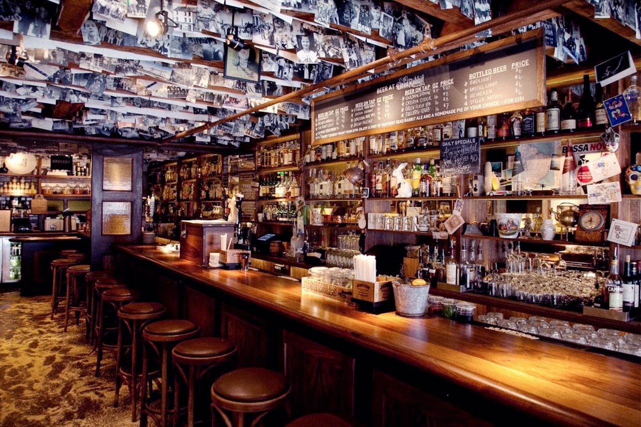 Interior of a traditional Irish pub with a bar lined with stools, decorated with an extensive array of photographs and memorabilia covering the ceiling and walls, showcasing a rustic wooden aesthetic and a well-stocked bar displaying various bottles and daily specials.







