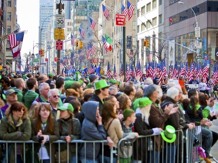 Crowd of people wearing green hats and celebrating St. Patrick's Day along a flag-lined street in New York City, with clear views of the parade and American and Irish flags in the background.