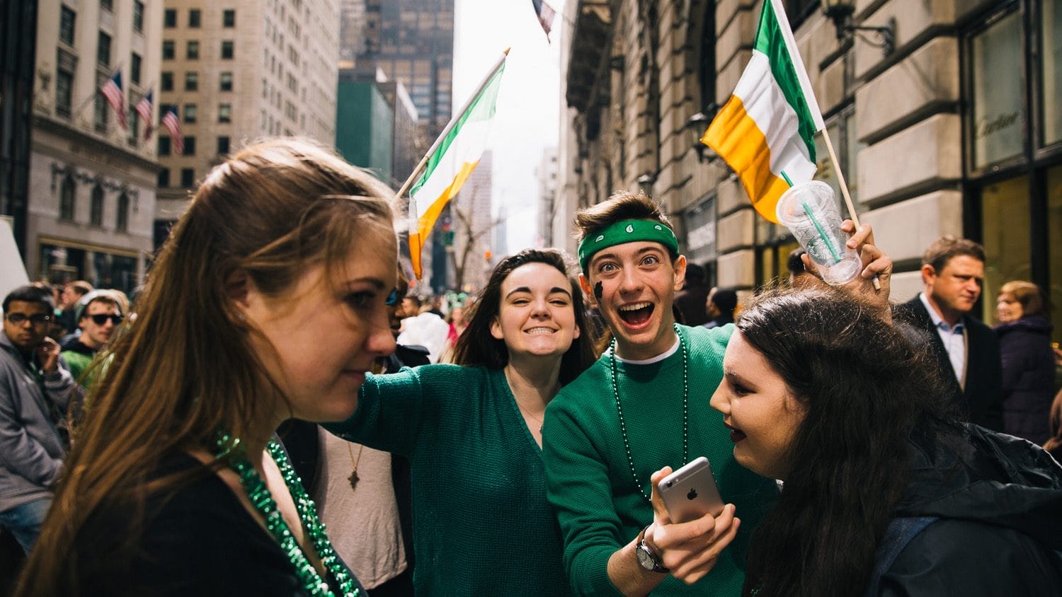 Young friends enjoying St. Patrick's Day celebrations in New York City, dressed in vibrant green attire and accessories. They are engaging with each other and sharing their excitement, capturing memories on a smartphone, surrounded by a crowd and Irish flags.