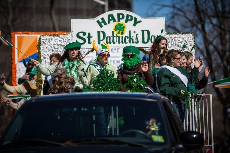 Participants on a parade float celebrate St. Patrick's Day, decked out in green outfits and accessories, waving to the crowd. The float is adorned with a "Happy St. Patrick's Day" banner and festive decorations, capturing the lively spirit of the holiday.