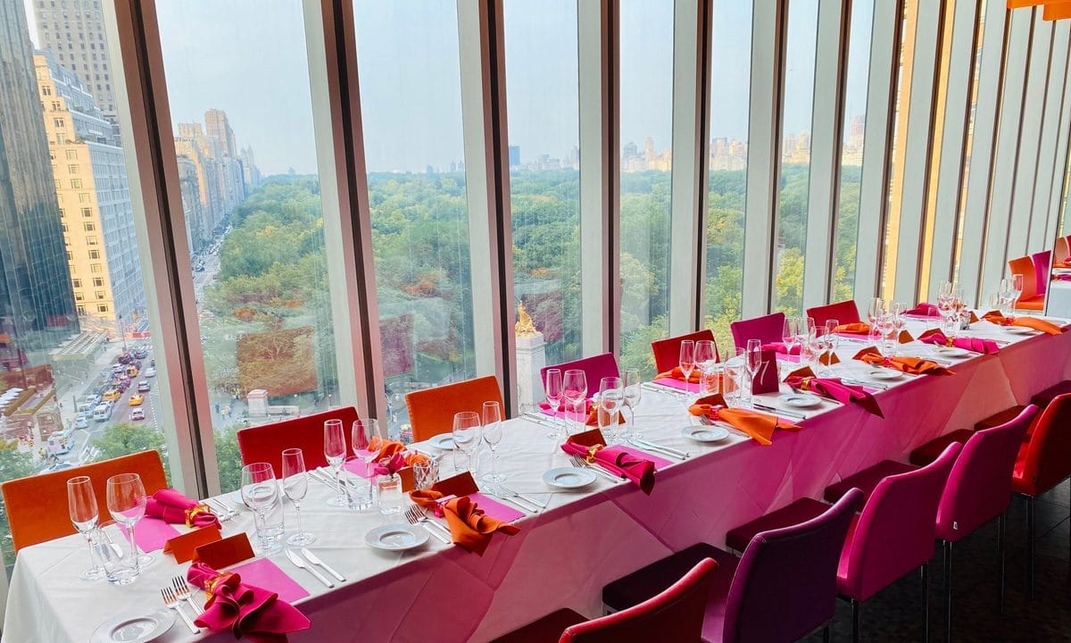 Brunch spot near Central Park with panoramic floor-to-ceiling windows offering sweeping park views. The table is elegantly set with bright napkins and glassware, creating a vibrant yet refined dining atmosphere.