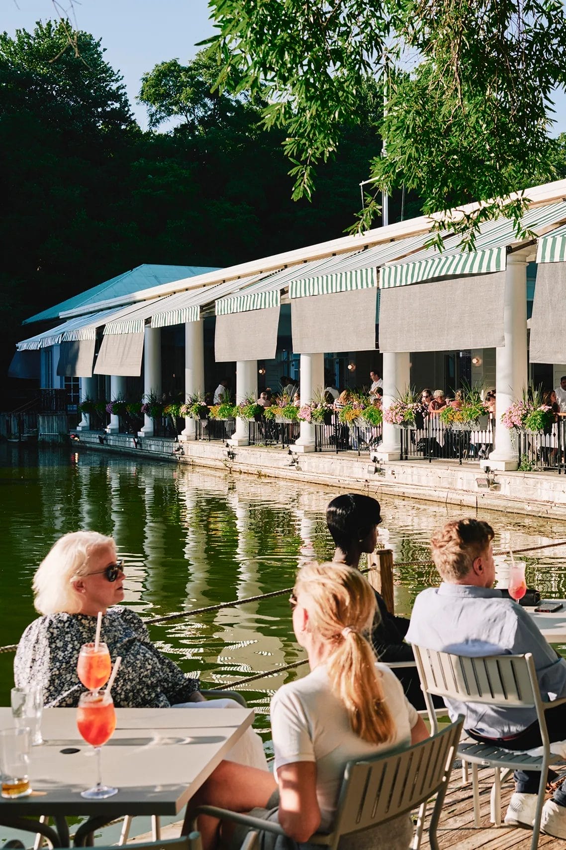 Lakeside brunch spot near Central Park featuring outdoor seating with scenic water views. Guests enjoy refreshing cocktails under the shade of trees, while the elegant terrace dining area is adorned with flowers and striped awnings.