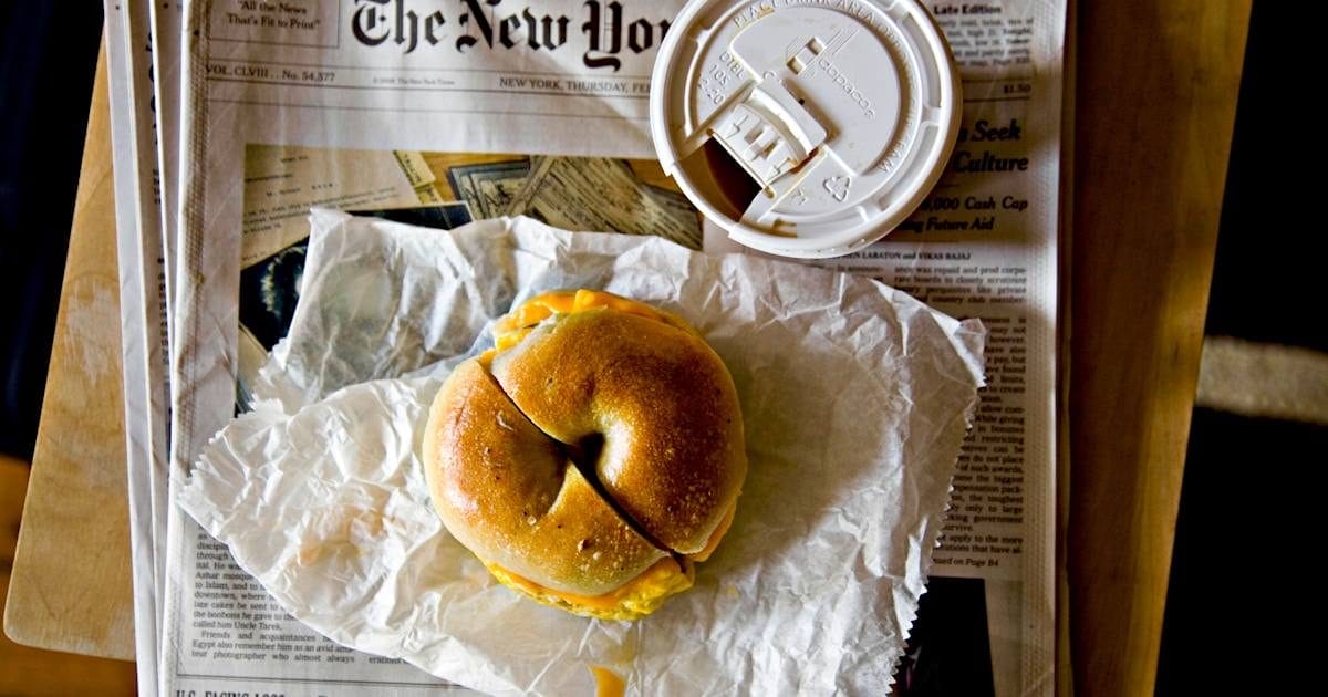 A fresh bagel sandwich on a New York Times newspaper with a takeaway coffee cup, capturing a typical NYC breakfast scene