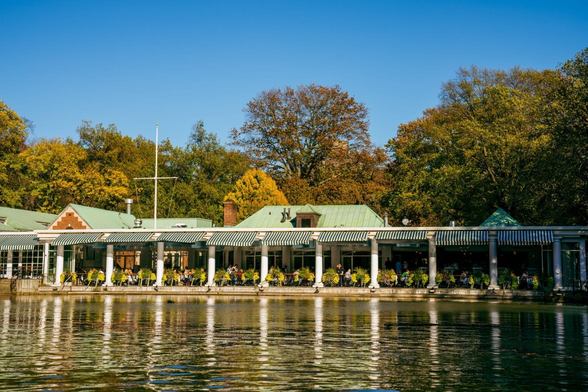 Scenic romantic restaurant near Central Park with lakeside outdoor seating, surrounded by lush trees and classic striped awnings, offering a serene dining experience.







