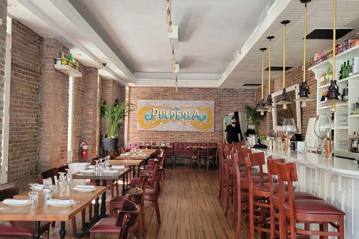 Interior of La Pulperia restaurant in New York, featuring a rustic ambiance with exposed brick walls and a long bar lined with high stools. The dining area is furnished with wooden tables and leather chairs, complemented by decorative pendant lights and vibrant wall art that adds a touch of Latin flair.