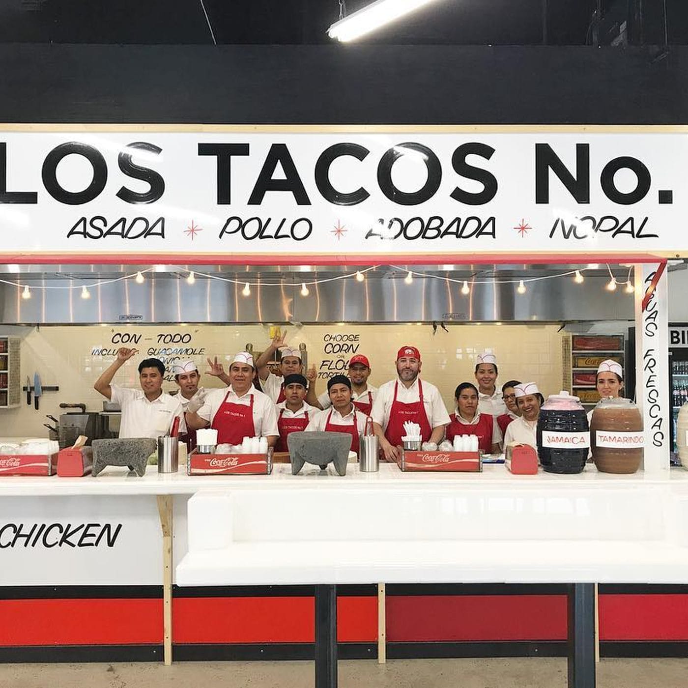 Staff at Los Tacos No. 1 standing proudly in their taco stand, with a sign listing their popular taco options like Asada and Pollo, creating a welcoming atmosphere for taco lovers.
