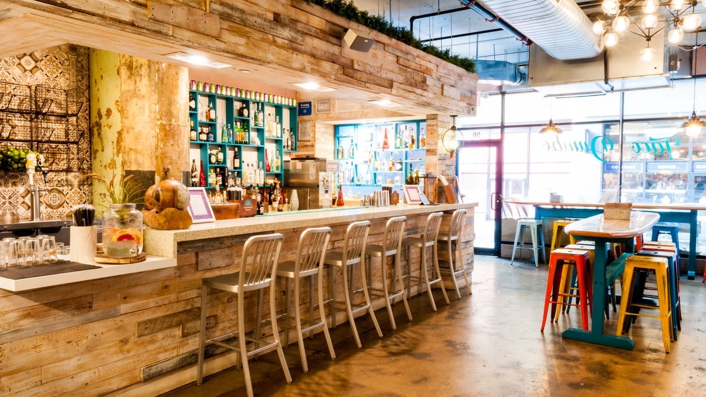 Chic bar area in a modern taco restaurant, featuring eclectic decor with reclaimed wood finishes, colorful bar stools, and a well-stocked backlit bar, creating a warm and inviting space for casual dining.
