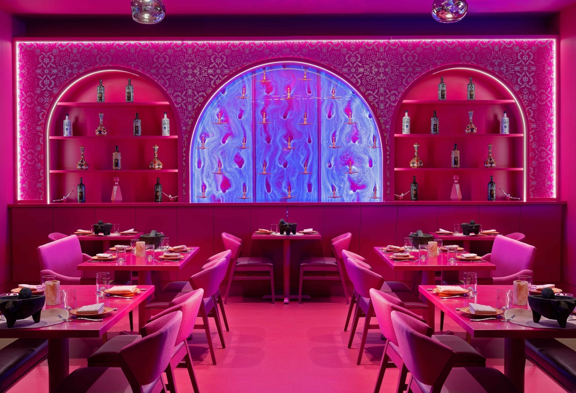 Vibrant and modern restaurant interior dominated by shades of pink, from the pink upholstered chairs to the vibrant pink walls. A striking blue and pink mural creates a dynamic backdrop, complemented by stylish shelving displaying eclectic decorative items and soft, glowing pendant lights that add a warm ambiance to the space.