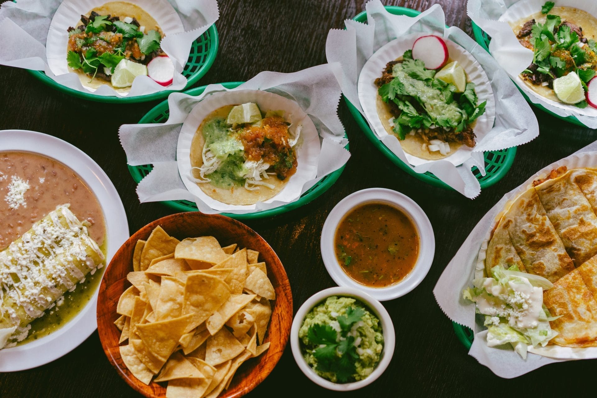 Assorted Mexican dishes spread on a dark wooden table, including fresh tacos with vibrant toppings, crispy tortilla chips, creamy guacamole, and rich sides of beans and quesadillas, offering a colorful and appetizing meal setup.