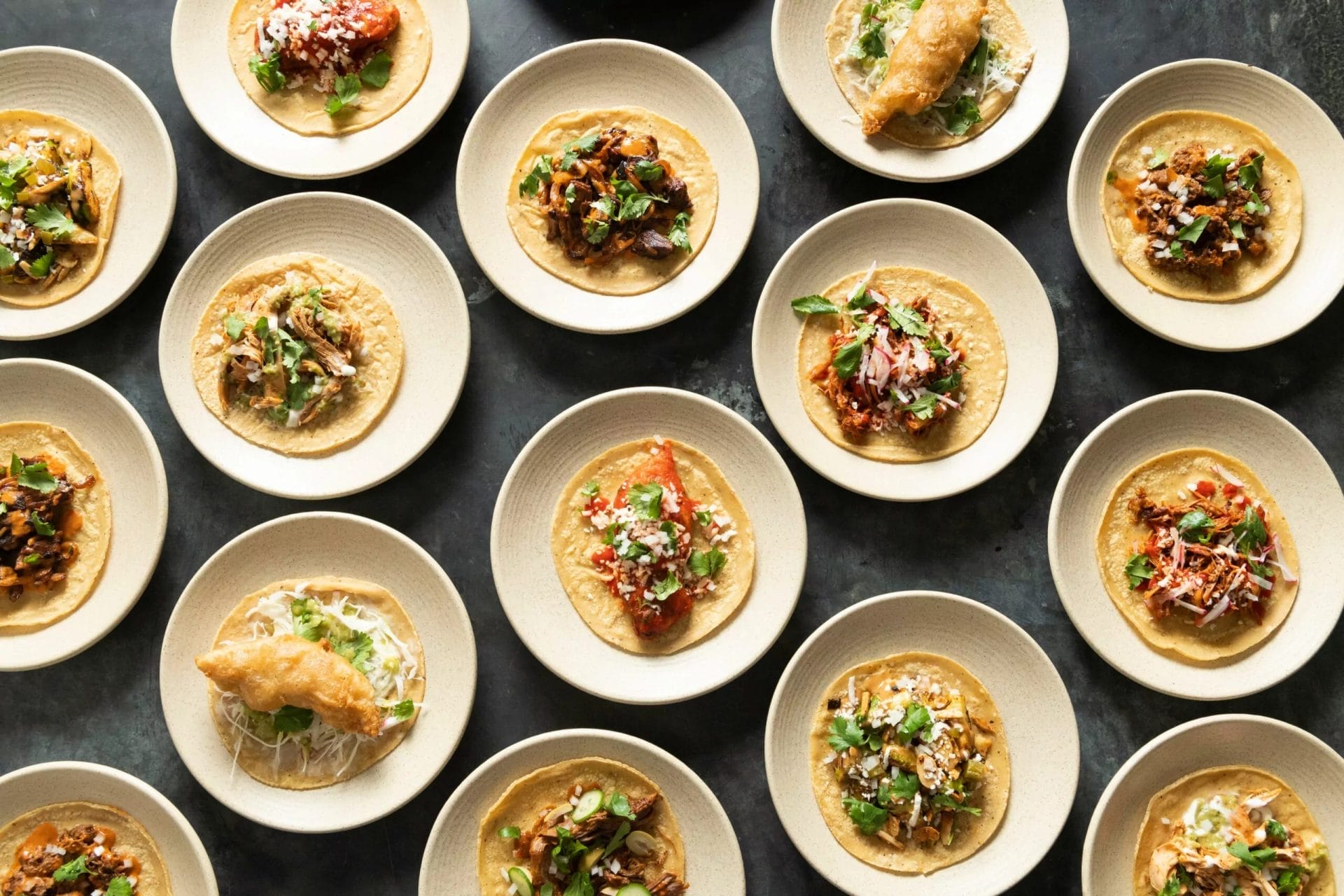 An array of gourmet tacos on individual plates, each beautifully presented with a variety of toppings and sauces on a dark stone countertop, showing the artful preparation and variety in modern taco cuisine.