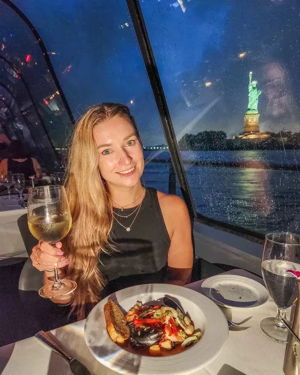 A woman enjoys a glass of wine, smiling in front of a delicious plate of food with the Statue of Liberty in the background.