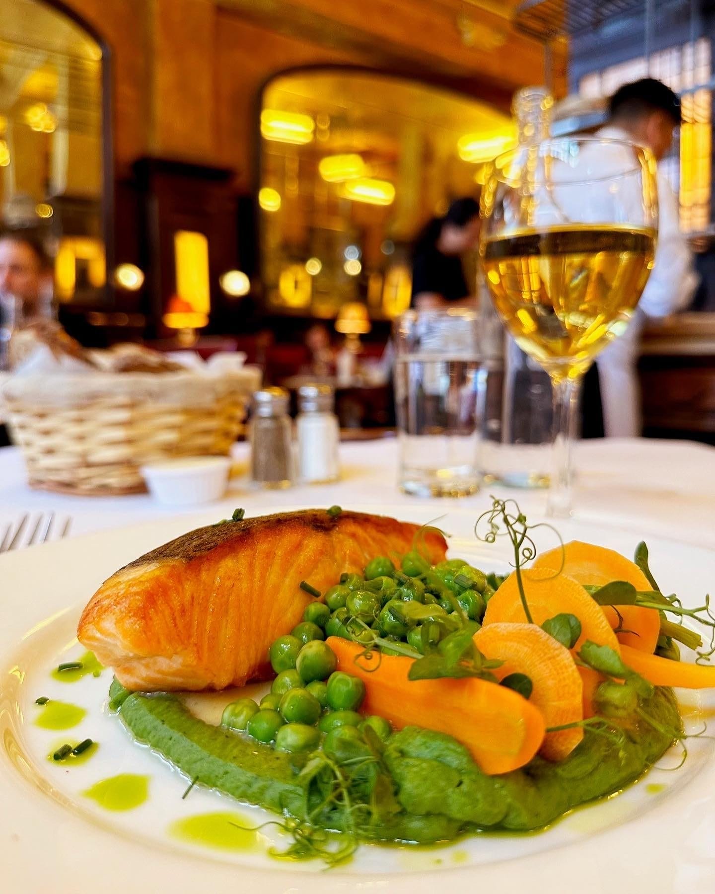 A plated dish featuring a seared salmon fillet with a golden crust, served on a bed of green pea purée. The dish is garnished with fresh green peas, thinly sliced carrots, and microgreens, with a drizzle of oil. A glass of white wine sits beside the plate in an elegant restaurant setting with warm lighting and a softly blurred background of diners and staff. It's open on Easter Sunday in New York City.