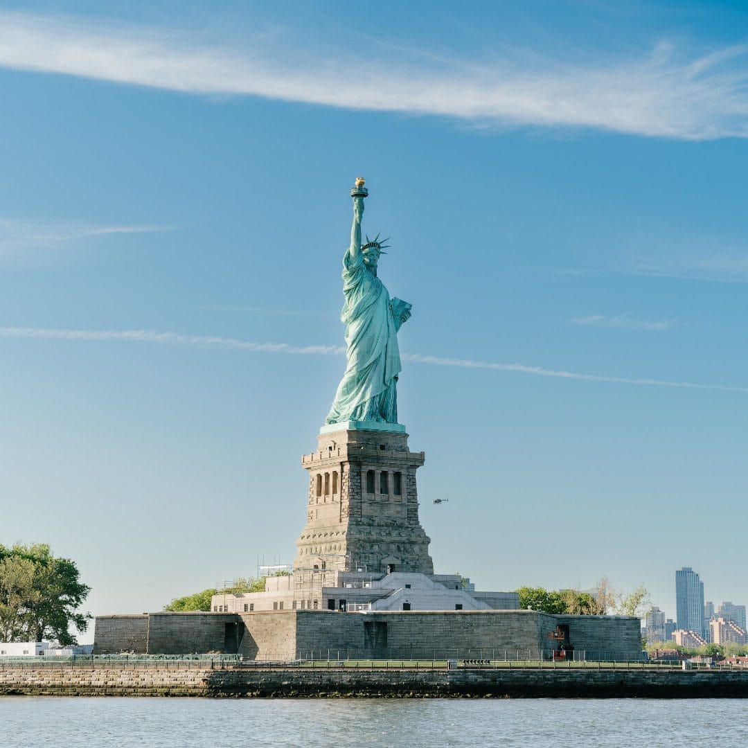 The Statue of Liberty stands tall against a clear blue sky, with its torch raised high. The surrounding area of Ellis Island and the distant city skyline are visible in the background. It's open on Easter Sunday in New York City.