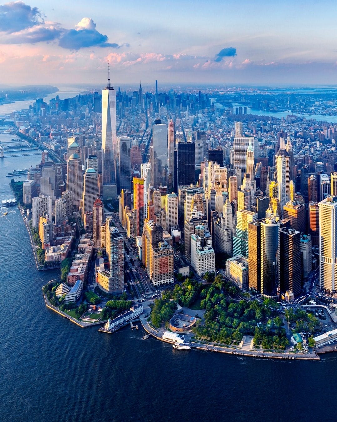 This is an aerial view of Lower Manhattan in New York City, featuring the One World Observatory at the top of One World Trade Center. The image captures the city's skyline with skyscrapers illuminated by sunlight, the waterfront of Battery Park, and the Hudson River in the background.