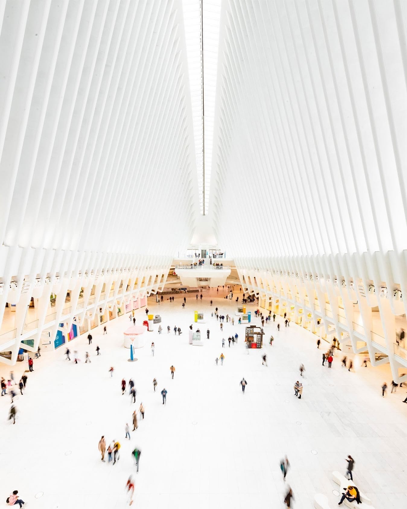 This is a high-angle view of the Oculus at the World Trade Center in New York City. The image showcases the bright, white interior with soaring rib-like architectural beams, a spacious central hall, and people moving throughout the space.