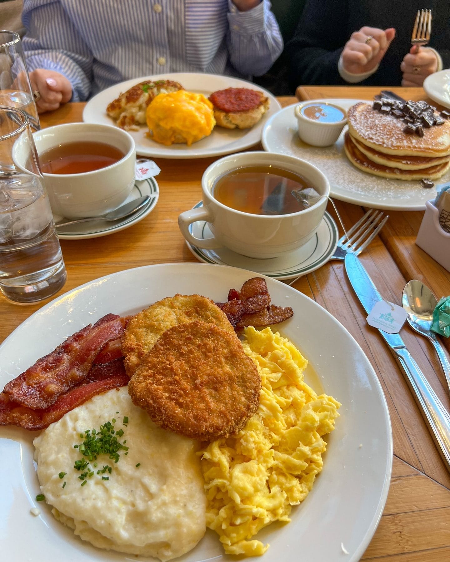Clinton St. Baking Co. features a plate of scrambled eggs, crispy bacon, hash browns, and creamy grits. In the background, other dishes include pancakes topped with chocolate chunks, tea, and eggs Benedict-style dishes, all served in a cozy, casual setting.