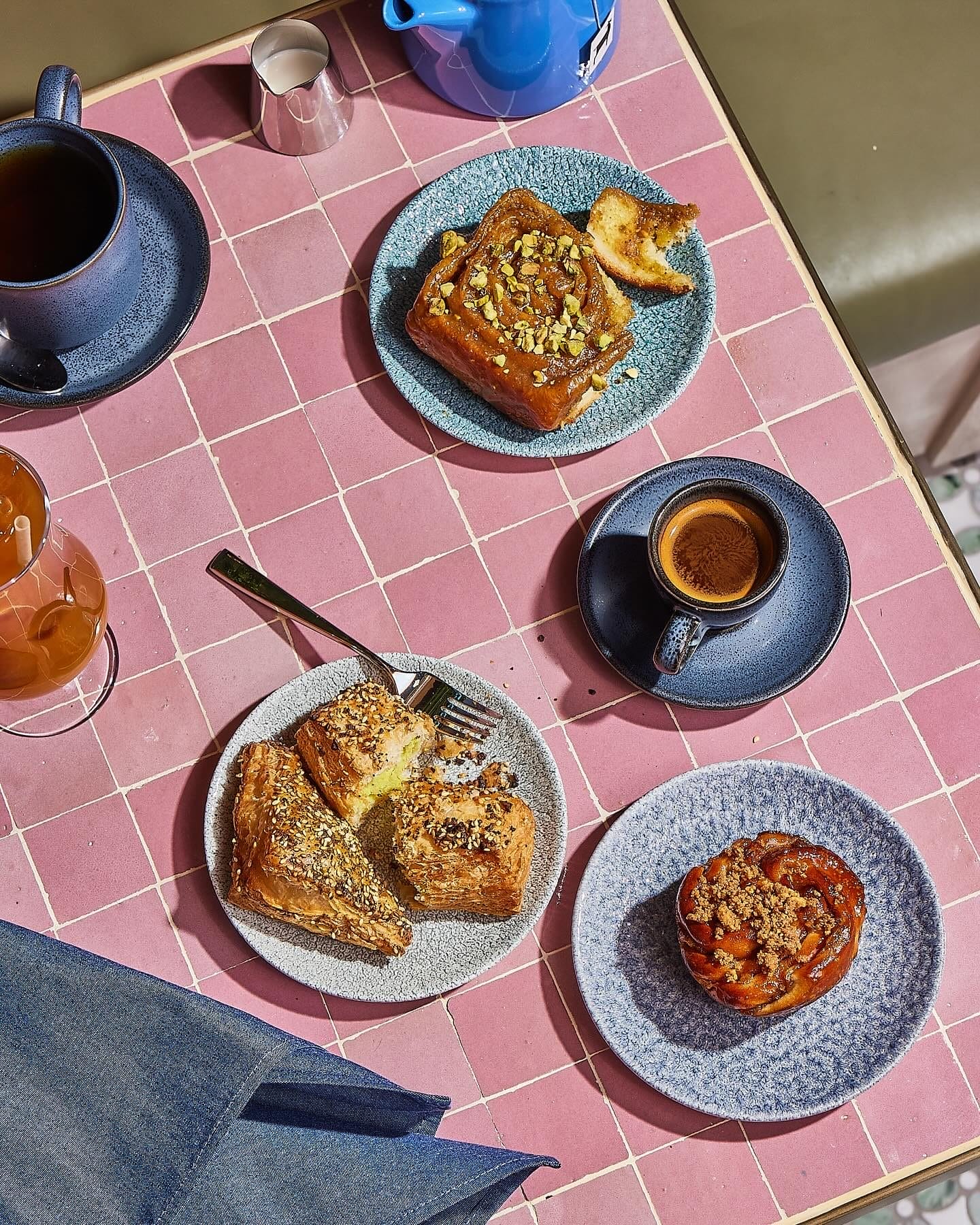 A vibrant brunch spread at K’Far Brooklyn featuring Israeli-inspired pastries, including a pistachio-topped sticky bun, flaky everything-seasoned borekas, and a caramelized pastry, served with coffee and tea on a pink-tiled table. A budget-friendly Easter brunch option with a unique twist on classic flavors.