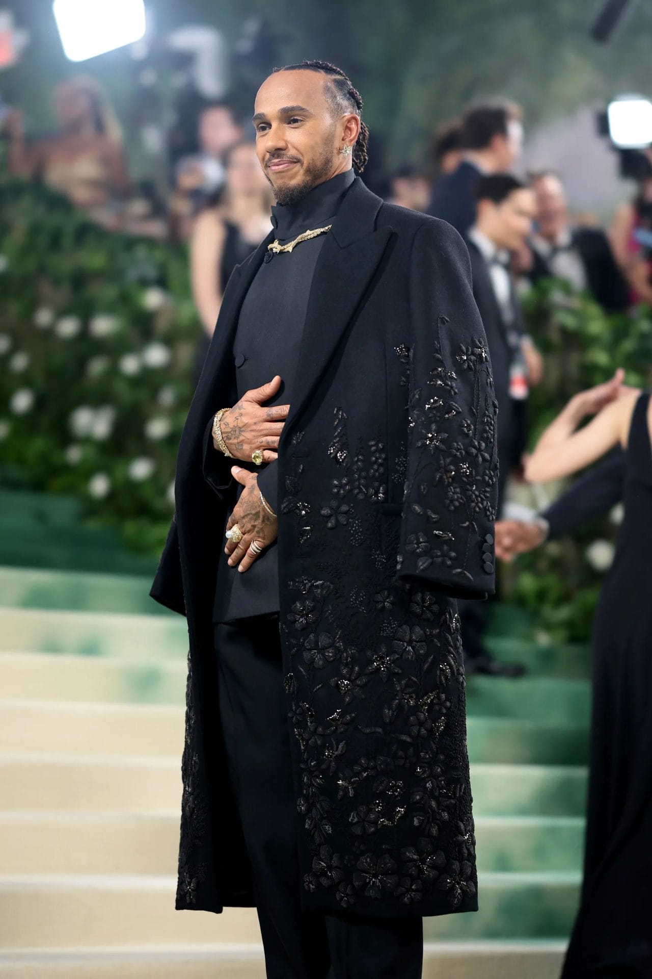 Lewis Hamilton poses at the Met Gala wearing an embroidered black overcoat layered over a dark ensemble with gold jewelry. Image from Vogue.com for the blog post "Met Gala 2025: Celebrities, Red Carpet, Theme & More."