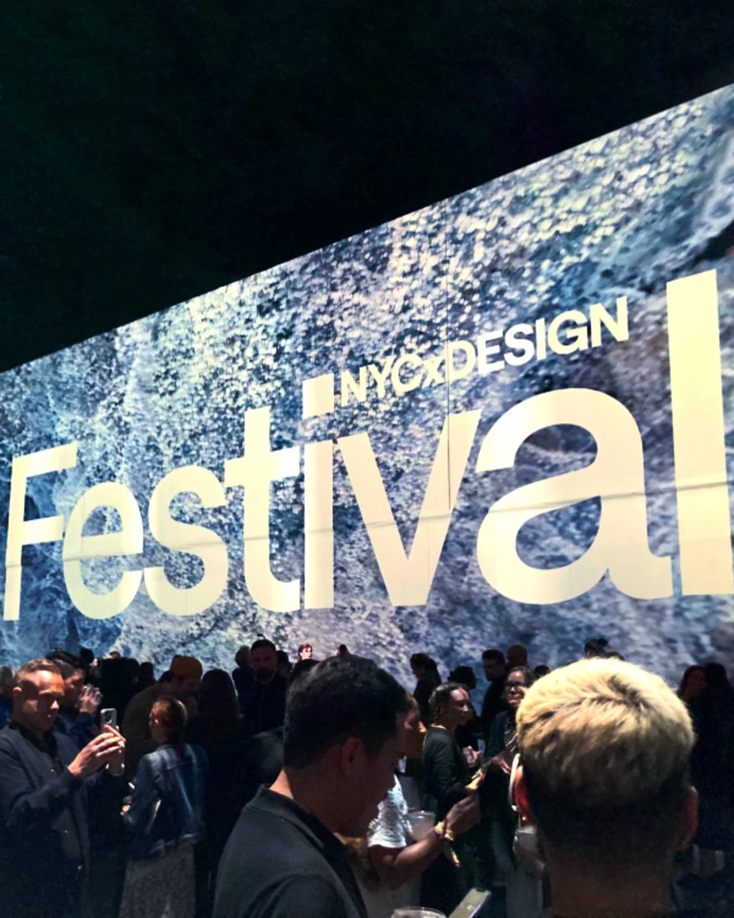 A crowd gathers at an NYCxDESIGN Festival event in front of a large illuminated wall displaying the word “NYCxDesign Festival.” This is for the blog post "Fun Things To Do in New York This May 2025."