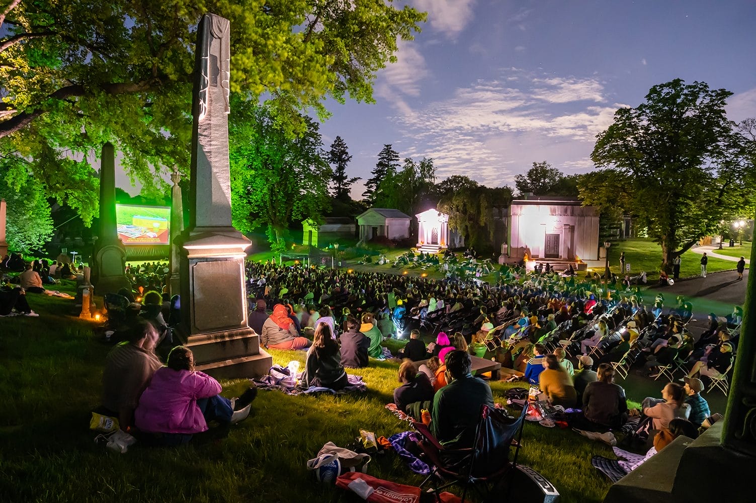 A large crowd watches an outdoor film screening at night in Green-Wood Cemetery, surrounded by tombstones and illuminated trees. This image is from Rooftop Films, part of the blog post "Fun Things To Do in New York This May 2025."