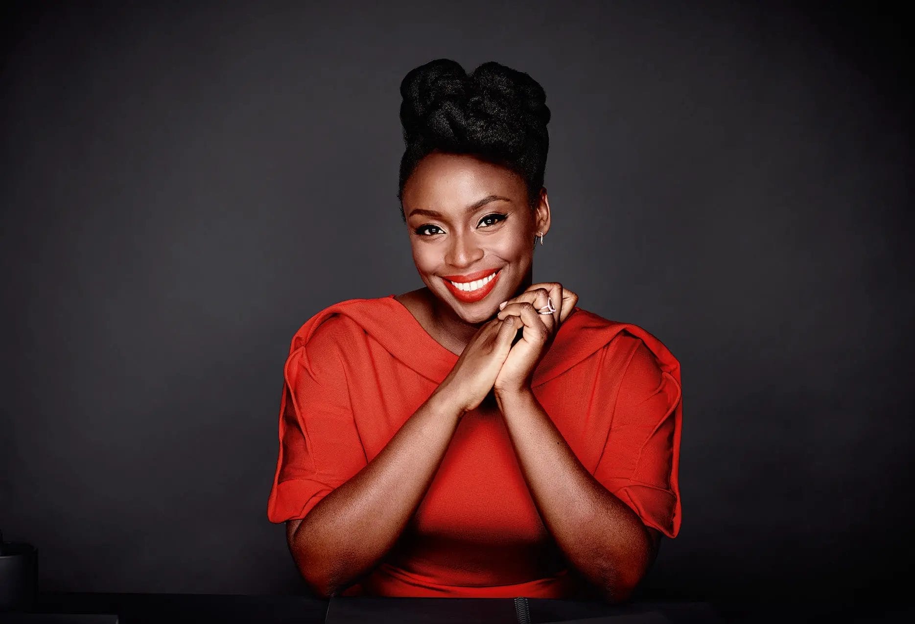 Chimamanda Ngozi Adichie poses against a dark background, smiling and wearing a red dress with her hands gently clasped.