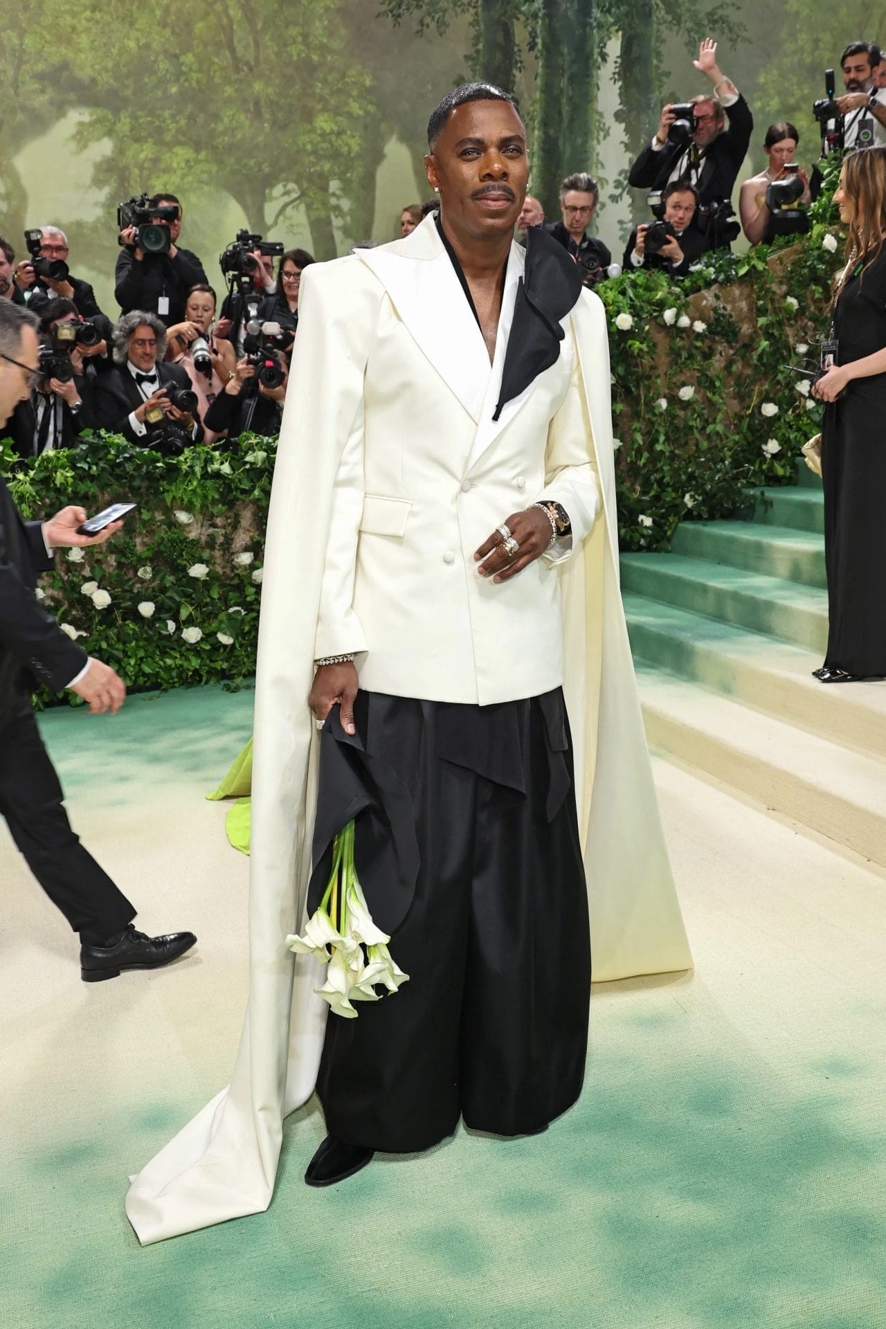 Colman Domingo on the Met Gala red carpet wearing a white cape tuxedo jacket with black trousers, holding a bouquet of calla lilies. Image from Vogue.com for the blog post "Met Gala 2025: Celebrities, Red Carpet, Theme & More"